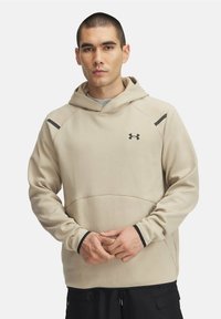 Beige huppari, jossa on kiristysnyörit huuvassa, mustat yksityiskohdat ja Under Armour -logo rinnassa. Pehmeä rakenne ja rento istuvuus.