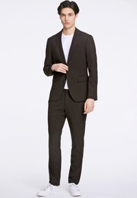 Bruin colbertjasje met bijpassende broek, gecombineerd met een wit T-shirt en witte sneakers. Gladde stof, getailleerde pasvorm en sluiting met een enkele knoop.