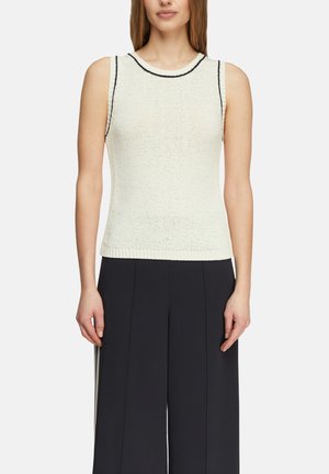 Kobieta ubrana w białą bezrękawnikową dzianinową bluzkę z czarną lamówką i czarne szerokie spodnie na gładkim tle.