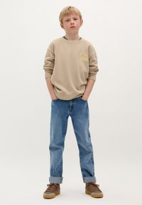 Beżowy sweter z żółtym nadrukiem na klatce piersiowej, zestawiony z niebieskimi dżinsami i brązowymi sneakersami. Luźny krój, widoczne detale mankietów na dżinsach.