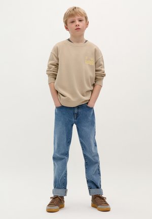 Beżowy sweter z żółtym nadrukiem na klatce piersiowej, zestawiony z niebieskimi dżinsami i brązowymi sneakersami. Luźny krój, widoczne detale mankietów na dżinsach.