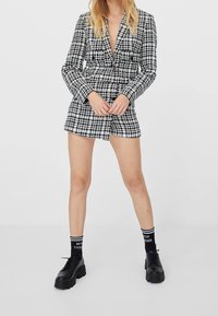 Kvinna i en svart och vit rutig kavajklänning med svarta plattformsskor och svarta strumpor som säger "Bättre tillsammans."