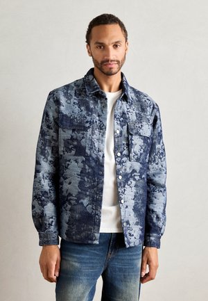 Denim jacket in blue and light blue tones, features a textured floral pattern, button-up front, and two chest pockets. Worn over a white shirt.