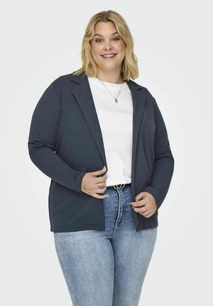 Blazer bleu marine foncé avec une coupe décontractée, des manches longues, des revers crantés et des poches avant, porté sur un haut blanc et un jean bleu clair.