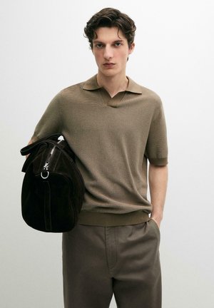Jeune homme portant un polo marron texturé à manches courtes et un pantalon assorti, portant un grand sac de sport en daim noir sur l'épaule.