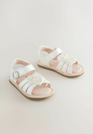 Valged rihmikuga sandaalid, varba peal südamekujuline sädelev dekoratsioon ja tekstuuriga roosa tallalabidas. Sulgemiseks külgkinnitus.