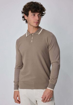 Jeune homme portant un polo beige texturé à manches longues avec une bordure blanche, regardant vers sa gauche, debout devant un fond uni.