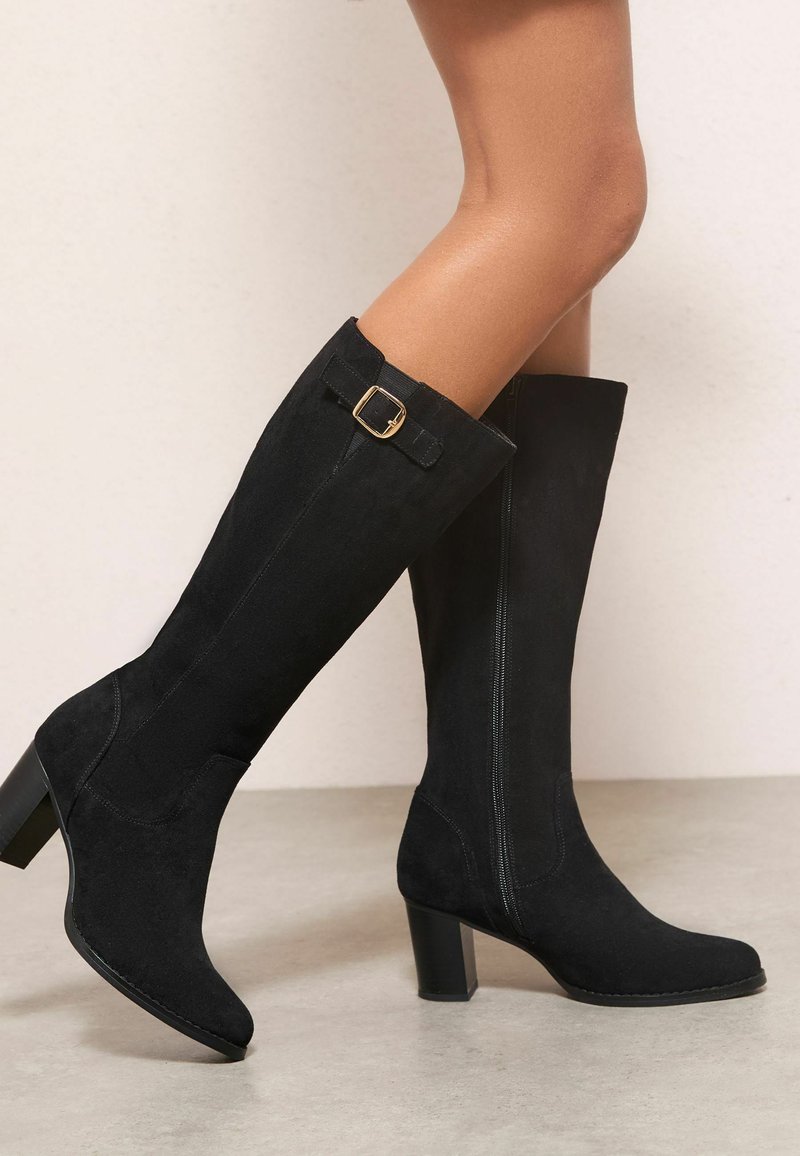 Bottes noires en daim montantes avec un bout carré, un talon bloc et un détail de boucle décoratif en haut. Texture lisse, fermeture éclair sur le côté.