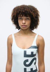 Femme aux cheveux bouclés portant un maillot de bain blanc avec de grandes lettres noires, se tenant devant un fond blanc uni, regardant vers l'avant.