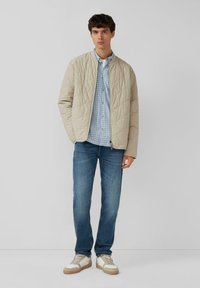 Beige gewatteerde jas met een ronde hals, gedragen over een lichtblauw geruit shirt; gecombineerd met blauwe jeans en meerkleurige sneakers.