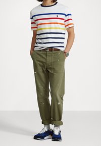 Camiseta de algodón a rayas con rayas azules, rojas y amarillas; pantalones verde oliva con manchas de pintura; zapatillas azules con detalles en blanco.