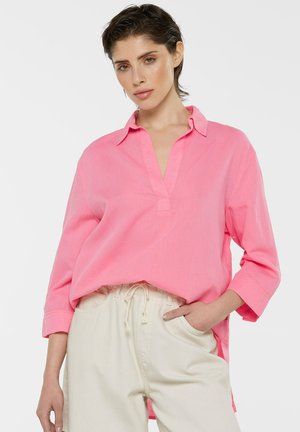 Pink relaxed-fit shirt with a notch collar, long sleeves, and a side-slit hem, paired with light beige drawstring pants. Smooth texture.
