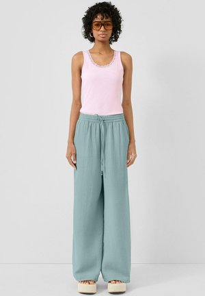 Femme aux cheveux bouclés et lunettes de soleil ambrées, portant un haut sans manches rose, un pantalon large bleu clair et des sandales à plateforme.