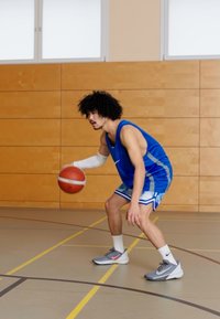 Mees korvpallur kannab sinist särki, valgeid varrukaid ja halli tossu roosa aktsendiga, palli dribbleerides spordisaali põrandal.