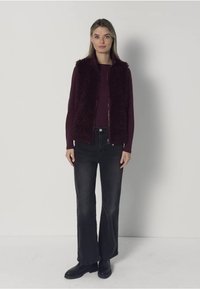 Bordeauxrode nep bontvest over een aansluitende top, gecombineerd met zwarte uitlopende broek en enkellaarsjes. Eenvoudig ontwerp met ritssluiting, zonder patronen.