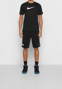 Černé tričko s kulatým výstřihem Nike s bílým logem, spárované s černými šortkami s bílým brandingem a černými teniskami s modrými akcenty.