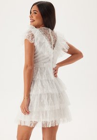 Abito bianco in pizzo con maniche a volant e gonna a strati. Presenta una scollatura a V sul retro e una texture di pizzo intricata. Design fino al ginocchio.