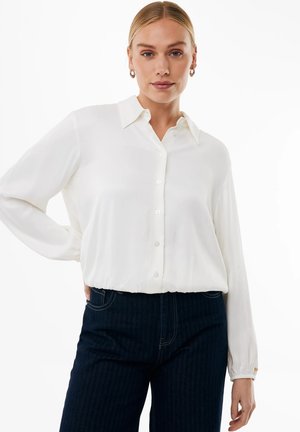 Kvinde med blondt hår iført en hvid knapbluse og mørke jeans, der står med den ene hånd på hoften mod en hvid baggrund.