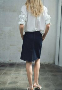 Witte gestreepte blouse met opgerolde mouwen, gecombineerd met een donkerdenim rok. De benen van een vrouw zichtbaar, met witte sandalen. Neutrale achtergrond.