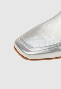 Silberne metallische Knöchelstiefel mit quadratischer Zehenform, glatter Textur und hellbrauner Sohle, die ein minimalistisches Design und eine elegante Verarbeitung betonen.