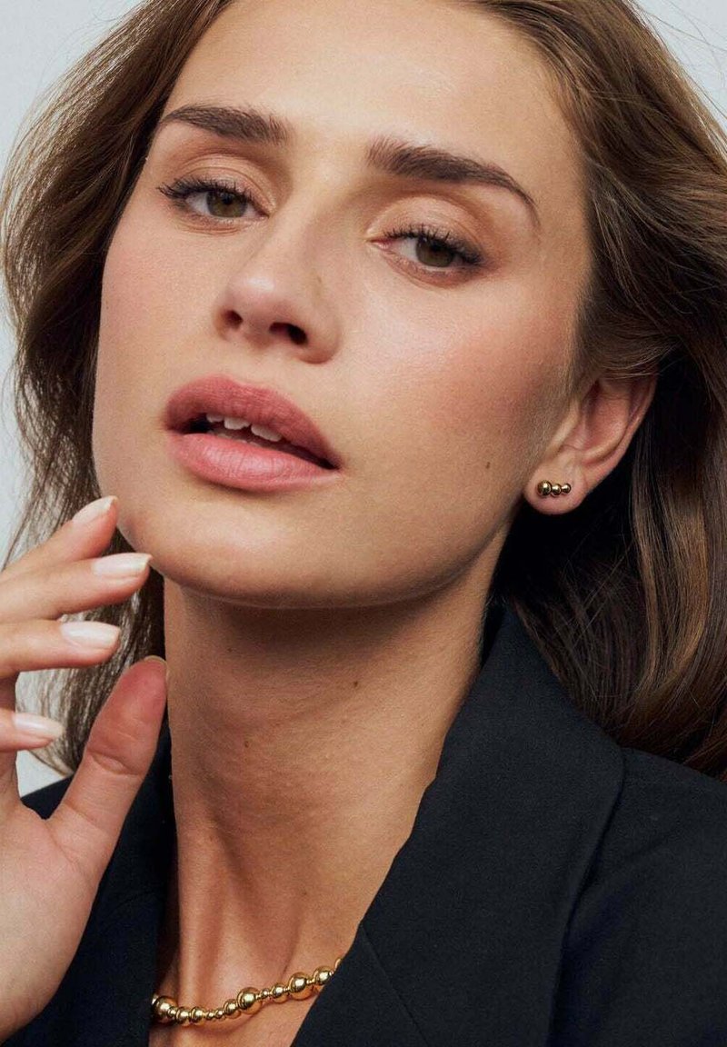 Guld studsörhängen har en slät, rundad design. Ett enkelt guldperlehalsband kompletterar looken mot en mörk svart jacka.