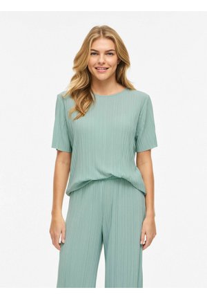 Žena s dlhými blond vlasmi má na sebe voľnú, svetlozelenú plisovanú blúzku s krátkym rukávom a zodpovedajúce široké nohavice, usmieva sa na bielom pozadí.