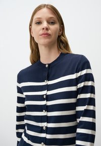 Cardigan a righe blu e bianche, con bottone frontale, scollatura rotonda, texture morbida, con righe orizzontali equidistanti e piccoli bottoni metallici.