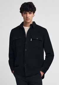 Czarna koszula z korduroju z długimi rękawami, dwoma kieszeniami na piersi i zapięciem na guziki. Posiada teksturowaną powierzchnię i kołnierz.