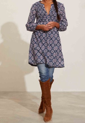 Mujer con túnica de manga larga azul con estampado, vaqueros azules y botas marrones de ante hasta la rodilla, de pie contra una pared lisa.