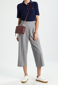 Donna che indossa una polo blu navy, pantaloni grigi a gamba larga e alla caviglia, sneakers bianche e una borsa a tracolla marrone.