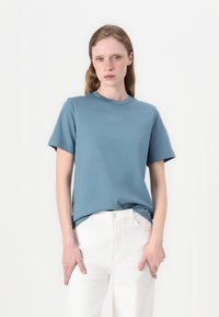 Mėlynas medvilninis marškinėliai su apvaliu iškirpimu ir trumpomis rankovėmis, ant priekinės dalies turi subtilų įspaustą Lacoste logotipą. Dėvimi su baltais džinsais.