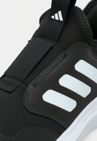 Primo piano di una sneaker Adidas nera con strisce bianche, linguetta nera e upper in tessuto testurizzato.