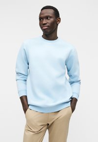 Ανοιχτό μπλε φούτερ με στρογγυλή λαιμόκοψη από μαλακό υλικό, με ριμπ μανσέτες και τελείωμα. Συνδυάζεται με μπεζ παντελόνια, προσφέροντας ένα casual στυλ.