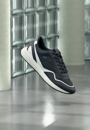 Μαύρο-άσπρο sneaker BOSS με υφή στη σόλα και σχέδιο από δίχτυ, αιωρούμενο πάνω από ένα γυαλισμένο δάπεδο από σκυρόδεμα σε μοντέρνο εσωτερικό χώρο.