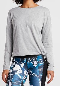 Grå långärmad topp med rund halsringning och justerbar sidoknytt. Kombinerad med svarta leggings med ett blått och vitt blommönster.