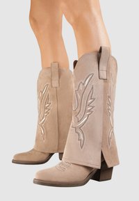 Bottes de cowboy en suède beige avec broderies décoratives en forme d'aile, bout carré et petit talon carré ; dotées d'une languette pour un enfilage facile.