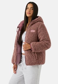 Różowa pluszowa kurtka z kapturem, z miękką, puszystą powierzchnią, zamkiem błyskawicznym i naszywką z logo na piersi. Gładka podszewka wewnętrzna.