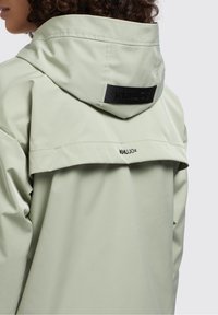 Jasnozielona nieprzemakalna kurtka z kapturem, z czarną naszywką z logo i warstwowym detalem na plecach. Gładka, matowa powierzchnia z minimalną ilością szwów.