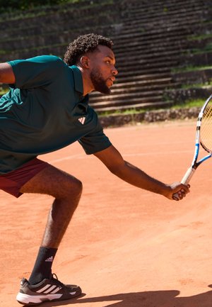 Athlete in a teal polo and maroon shorts lunging on a clay court, holding a tennis racket with a black and white sneaker visible.