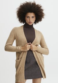 Beige gyapjú kardigán hosszú ujjak, nyitott elüls�ő rész és oldalsó zsebek jellemzik, barna garbó fölött viselve, szürke szoknyával párosítva.