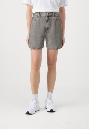 Person iført højtaljede, falmede grå denimshorts, hvid kort top, hvide sokker og hvide sportssko mod en ensfarvet baggrund.