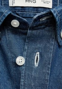 Denim košulja koja se sastoji od plave tkanine s teksturiranim završetkom, bijelim plastičnim dugmadi i malom dugmadi sa strganoj ivici.