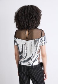 Korte mouwen top met zilveren pailletten en een zwarte doorschijnende achterkant, sleutelgatdetail en een losse fit, gecombineerd met gestreepte broek.