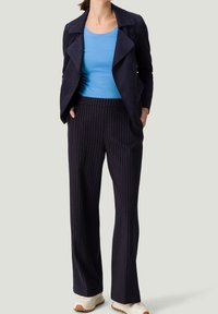 Nainen, joka on pukeutunut siniseen paitaan, mustiin raidoitettuihin housuihin, tummansiniseen avonaiseen takkiin ja valkoisiin tennareihin, seisoo kädet taskuissa.