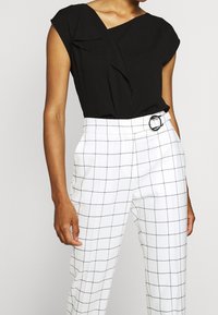 Haut noir à design en couches ; manches courtes. Pantalon blanc à motif quadrillé avec ceinture à boucle circulaire. Texture de tissu lisse.