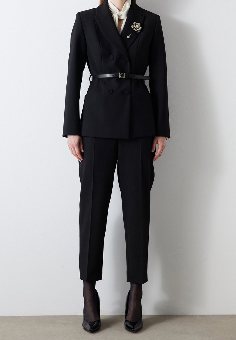 Personne portant un blazer noir ajusté à double boutonnage avec ceinture, pantalon noir court, collants noirs transparents, talons pointus noirs et col de chemise blanc.