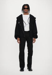 Jachetă neagră oversized cu fermoar, cu mâneci bufante, cămașă albă cu mâneci lungi și design grafic, pantaloni negri și ochelari de soare negri.