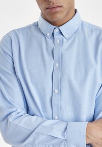 Camisa azul clara de botones, hecha de un tejido texturizado, con un collar clásico, mangas largas y botones blancos. Logo sutil en el pecho.