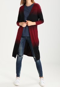 Lång cardigan i gradient nyanser av bourgogne och svart, tillverkad av mjukt material. Den har öppen framsida och åtsittande ärmar. Bärs över en blå topp.