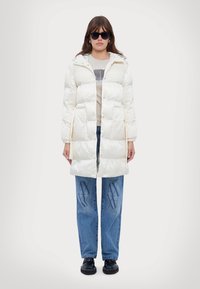Lang hvit pufferjakke med knappelukking, hette og teksturert glans. Kombinert med lyseblå jeans med mørkeblå grafikk og svarte sko.